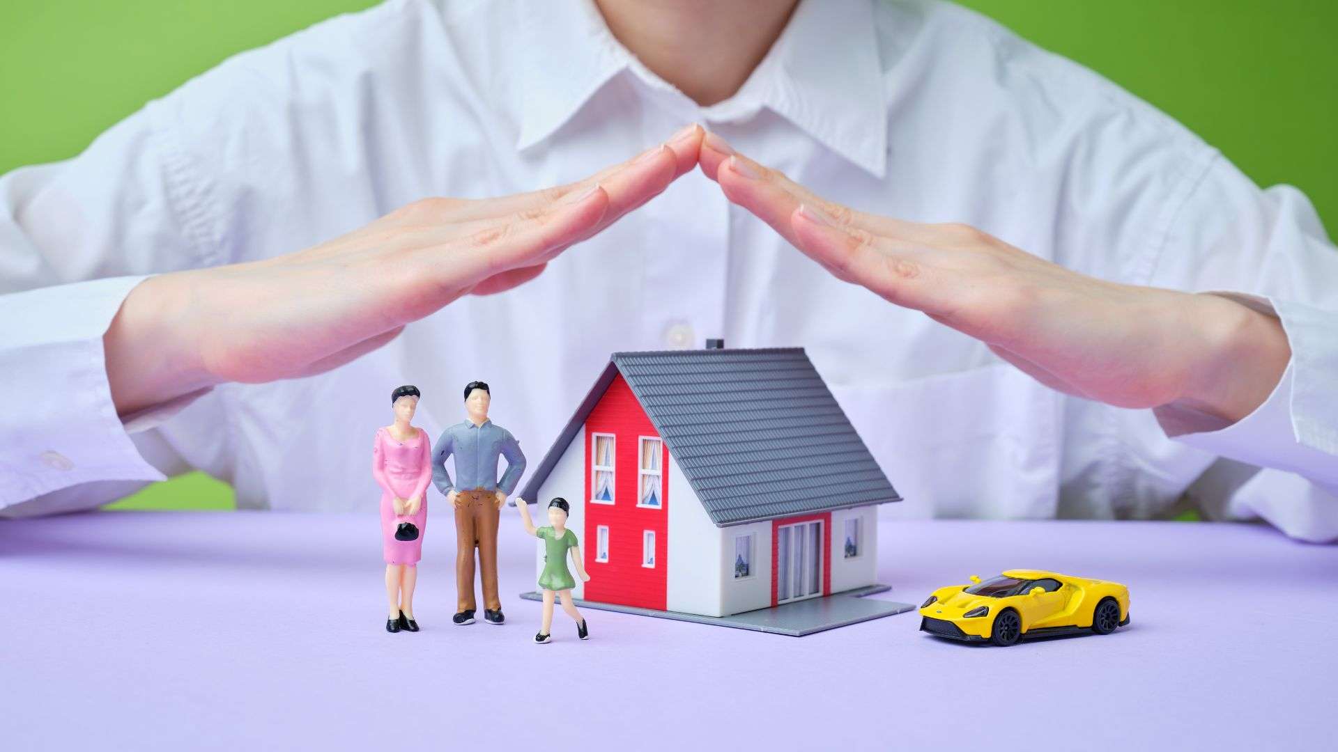 A person's hand is placed above miniature figures of a family and a house, symbolizing protection and insurance.
